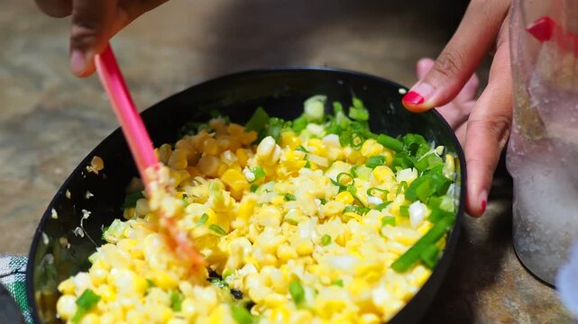 Close-up of hands mixing fresh corn and green onions in a black bowl with a pink spatula