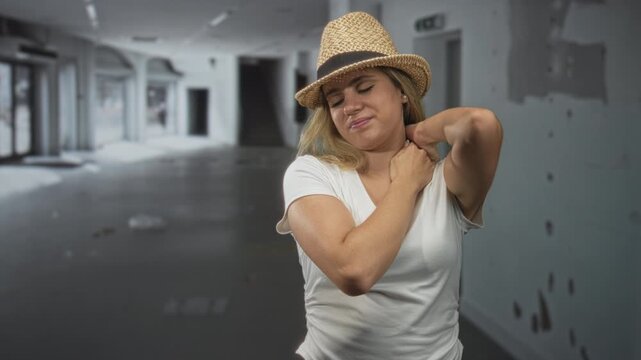 Woman touching her neck and shoulder in an empty building wearing straw hat and white tshirt, expression of grimace; travel fatigue.