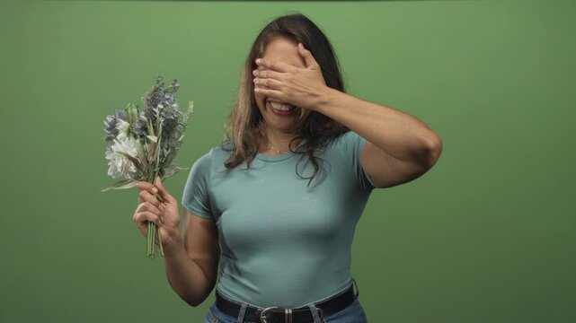 Woman holding bouquet of white and muted blooms, covering her eyes with one hand in green studio; shyness.