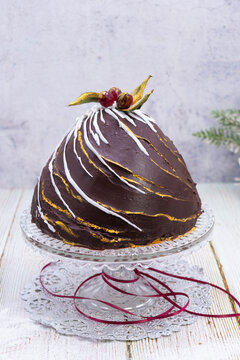 An elegant chocolate Christmas bombe cake with gold leaf accents, white chocolate drizzle, and festive sugar holly berry topper on a crystal cake stand.