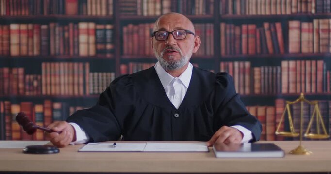 Judge holding gavel and talking verdict in courtroom with law library books. Senior man in judge robe sitting at wooden desk with justice scales to lead proceeding, court hearing of criminal case