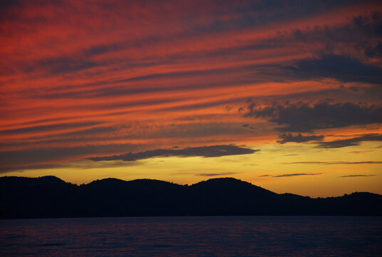 Colorful blue hour sunset over the Adriatic Sea with the dark silhouette of Ugljan Island on the horizon. Dramatic clouds painted with orange, pink and purple tones reflect on the calm water.