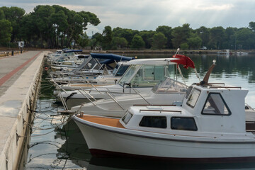 Row of white motorboats moored along stone embankment promenade with pine trees and cloudy sky in European coastal marina. Concept of nautical leisure, Mediterranean harbor life, summer boat tourism.