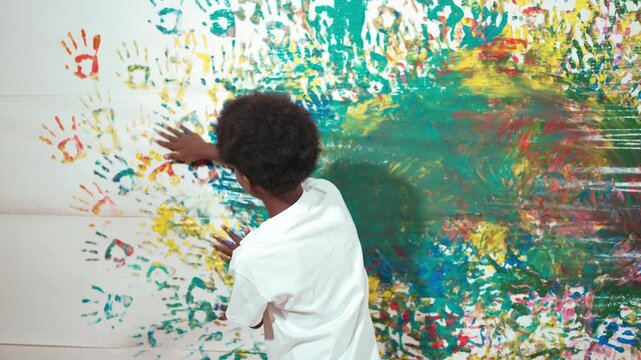 Back view of happy african boy painted the stained wall with colorful hand while wearing casual white shirt in art lesson.Smart student use hand print to make creative artwork. Education. Edification
