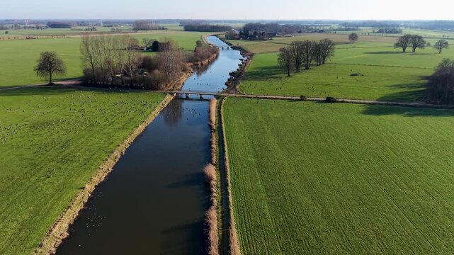 Aerial view of a rural landscape with a canal curving through green fields, two small road bridges crossing it, and a nearby house with outbuildings set among open farmland.