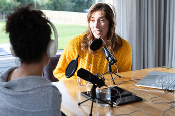 Two female podcasters recording at home studio table, host in mustard sweater using mics and laptop
