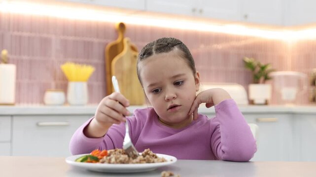 Unhappy little girl refusing to eat buckwheat and vegetables at light table in kitchen