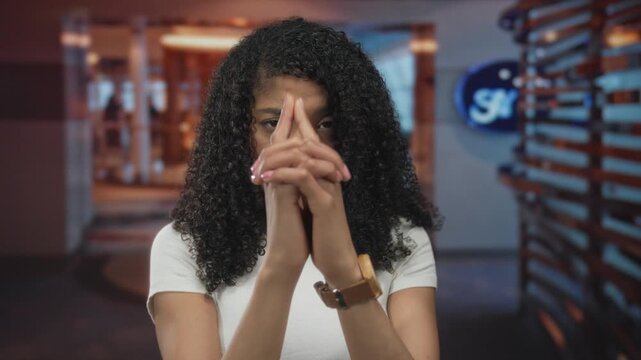 Woman holds interlaced fingers as pistol shape near temple inside hotel building, serious expression; tension.