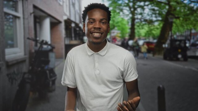 Man in white polo shirt claps hands and smiles broadly on street lined with buildings; anticipation eagerness.