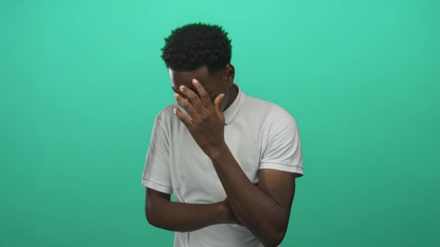 Young man covers face with hand in green studio; embarrassment vulnerability introspection selfconsciousness.