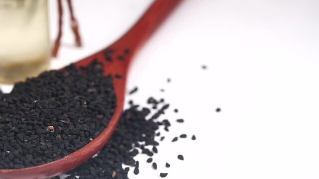 A close up cinematic view of black cumin seeds in a wooden spoon with a bottle of black seed oil in the background on white surface