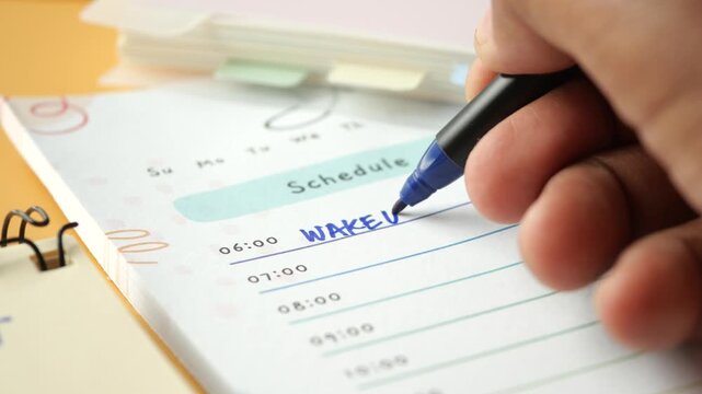 Person writing daily schedule in paper planner using blue ink pen for time management