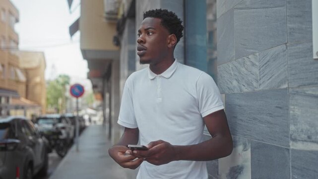 Man in white polo shirt holding smartphone scrolls screen while leaning against stone building on sunlit city street; contemplation.