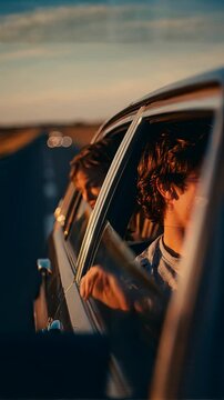 Two smiling young men lean out of car windows into the warm golden light of a sunset road trip, with bokeh highway lights and a broad open landscape glowing in the background.