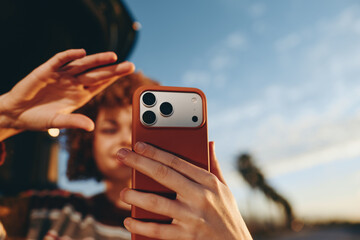Woman smiling while taking a selfie with smartphone outdoors. She wears a rainbow sweater and poses...