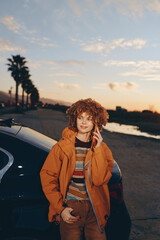 Fototapeta premium Young woman with curly hair smiling while talking on smartphone near car at sunset. She wears a rainbow sweater and orange jacket, lifestyle concept with warm natural light outdoors.