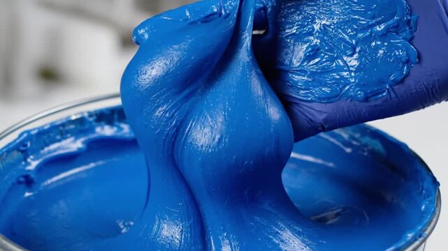 A gloved hand expertly manipulates a thick, vibrant blue substance, pulling it slowly from a container. The rich, viscous material stretches and folds, showcasing its unique texture and brilliant colo