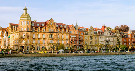 Fototapeta premium Historic buildings line the riverbank in a vibrant city during a sunny afternoon with clear skies and calm waters