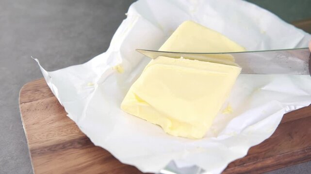Person cutting block of fresh butter with silver knife on wooden board in kitchen