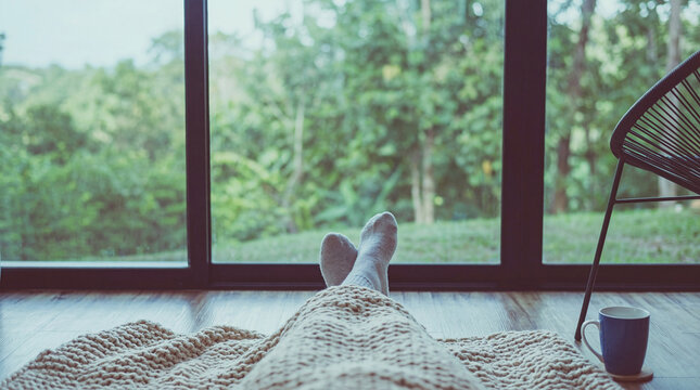 First-person view photograph of legs in cozy socks and a blanket, relaxing on the floor with a forest view.