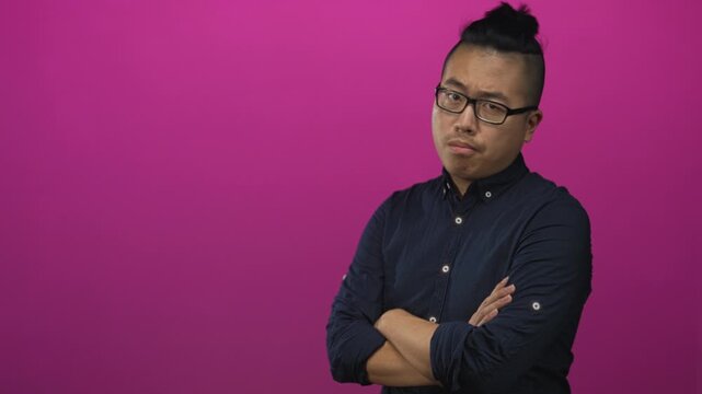 Young man with crossed arms and glasses in studio wearing navy shirt with rolled sleeves against pink backdrop; quiet confidence.