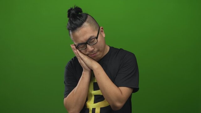 Young man wearing bitcoin t shirt and glasses, hands to cheek for sleep in studio with green backdrop; sleep calm.