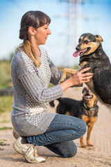 Obraz premium Woman kneeling on a dirt path interacts with a small black dog, surrounded by greenery and power lines under a clear blue sky