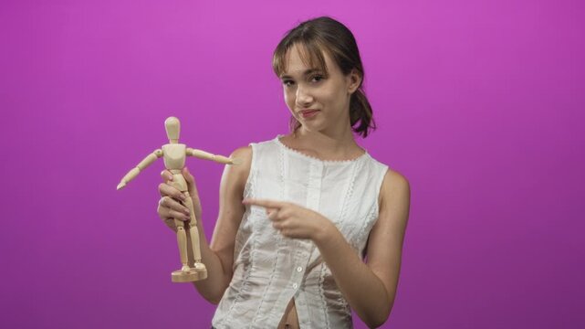 Young woman holding a wooden mannequin and pointing finger at the figure in a pink studio with magenta wall; playful curiosity.