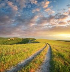 Rural landscape featuring a winding dirt road leading through lush green hills toward a vibrant sunset sky. A peaceful and atmospheric nature background. Travel, adventure, and freedom-themed designs.