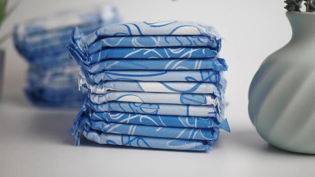 Stack of individually wrapped sanitary pads with blue floral pattern displayed on a white surface next to a vase