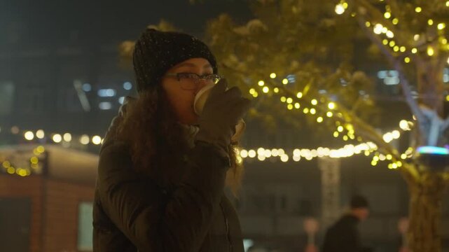 Woman With Glowing Drink In Market. Young Lady Smiles Amid Glowing Lights And Bustling Market Ambiance. Cheerful Woman Holds Warm Beverage Under String Lights In Lively Market Surroundings