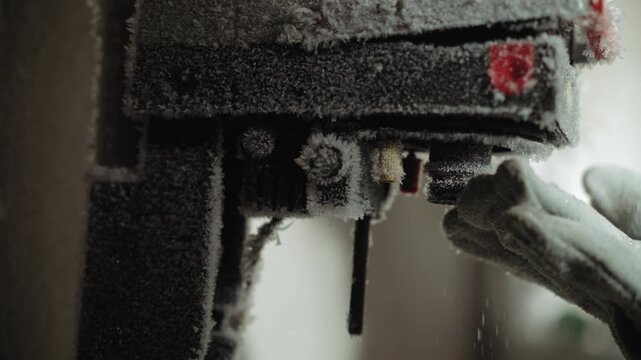 Gloved hands perform delicate repair on frozen drill press, removing ice and testing chuck, textured frost on housing, steady operator movements, gritty survival workshop mood after nuclear winter