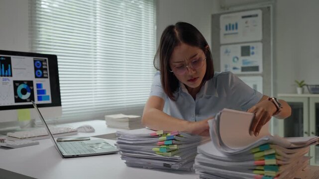 Stressed asian businesswoman searching for document in paperwork pile at office