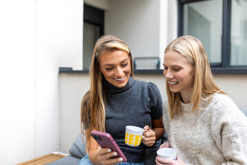 Fototapeta premium Two young female friends using smartphone on balcony. Communication, friendship, and digital lifestyle concept. Happy women looking at phone screen while holding coffee mugs. Social media app.