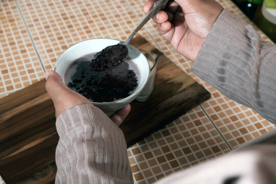 Woman's hand stirring a bowl of Bubur Ketan Hitam (black sticky rice), a sweet Indonesian porridge made from black sticky rice, coconut milk, and palm or cane sugar.  Indoensian traditional food. 