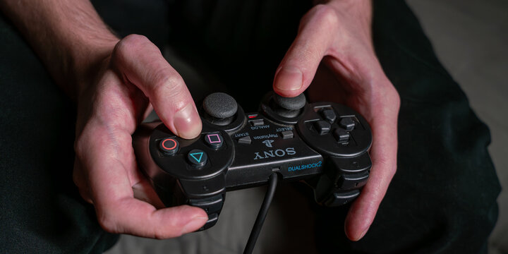 A close-up shot of a person's hands holding a black Sony DualShock2 controller for PlayStation 2 PS2 games console while playing a video game. The focus is on the controller and the hands.