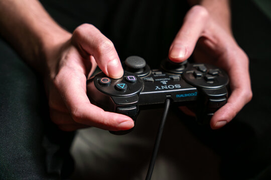 A close-up shot of a person's hands holding a black Sony DualShock2 controller for PlayStation 2 PS2 games console while playing a video game. The focus is on the controller and the hands.