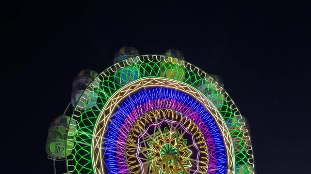 A brightly lit Ferris wheel glowing with multicolored lights in geometric star patterns against the dark night sky, creating a festive and vibrant atmosphere.