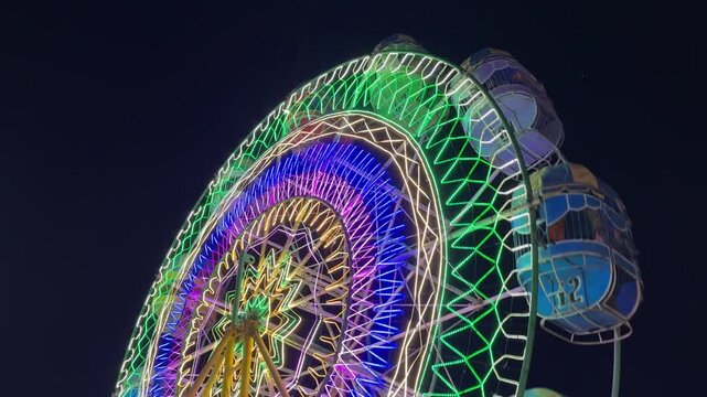A Ferris wheel glowing with multicolored lights in concentric circles and starburst patterns against the dark sky, creating a festive and lively atmosphere