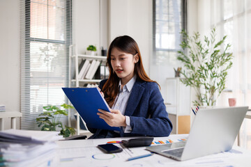 This image captures two women sitting side by side in a bright office space, involved in phone conversations while working on their laptops in a professional environment.
