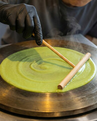 Detailed professional hand wearing black glove carefully spreading green matcha crepe batter on hot round griddle using wooden spreader tool with precision and focus on cooking process