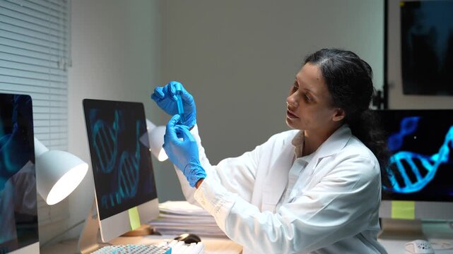 Focused woman in a white coat and gloves working late in a research laboratory, carefully examining a test tube sample with DNA helix structures displayed on computer screens in the background