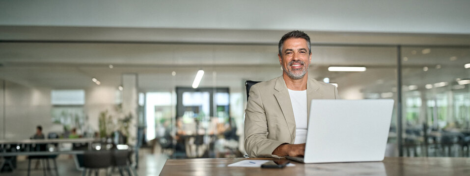 Happy mature bank manager looking at camera in office. Middle aged professional business man executive financial investor leader using laptop computer working sitting at desk. Portrait. Copy space.
