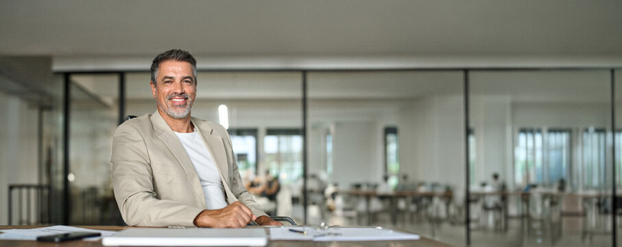 Happy middle aged business man ceo sitting at office desk. Smiling mature confident professional executive manager, 50 years old businessman leader wearing suit posing for corporate portrait at work.