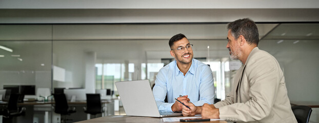 Young smiling professional latin business man working in corporate office with older executive manager using laptop technology having conversation on financial project strategy at work meeting.