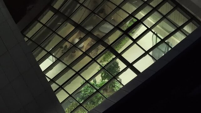Drone view of modern building glass roof revealing illuminated interior atrium with plants, seating area, and contemporary architecture at night in Vilnius, Lithuania.