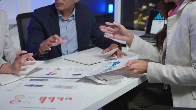 Group of dedicated asian business professionals analyzing graphs and charts during a late night meeting. Colleagues brainstorming over paperwork