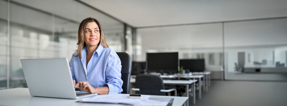 Happy mature middle aged professional business woman bank worker or hr manager, corporate executive looking away at work working on laptop computer in office thinking of business technology future.