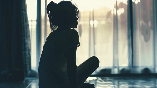 Silhouette of a child sits alone by window. Soft light filters through sheer curtains. Child's hair is tied loosely in a ponytail. Stillness fills the quiet room. Sunlight casts gentle glow on floor