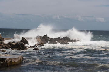 Fototapeta premium Powerful ocean waves crash against rugged rocks and a concrete breakwater on a cloudy day